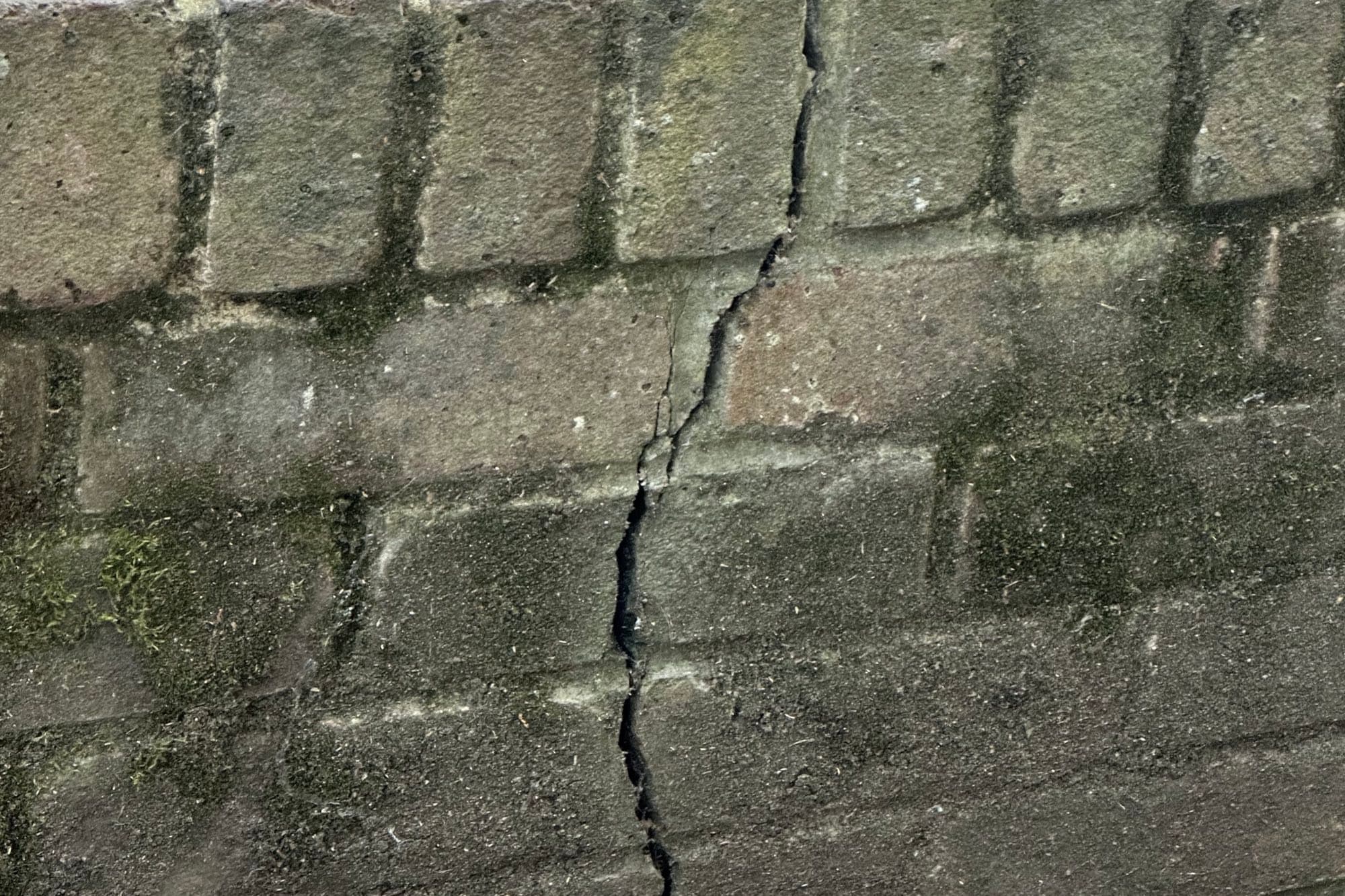 Close-up of a vertical crack running through a weathered, moss-stained concrete block retaining wall.