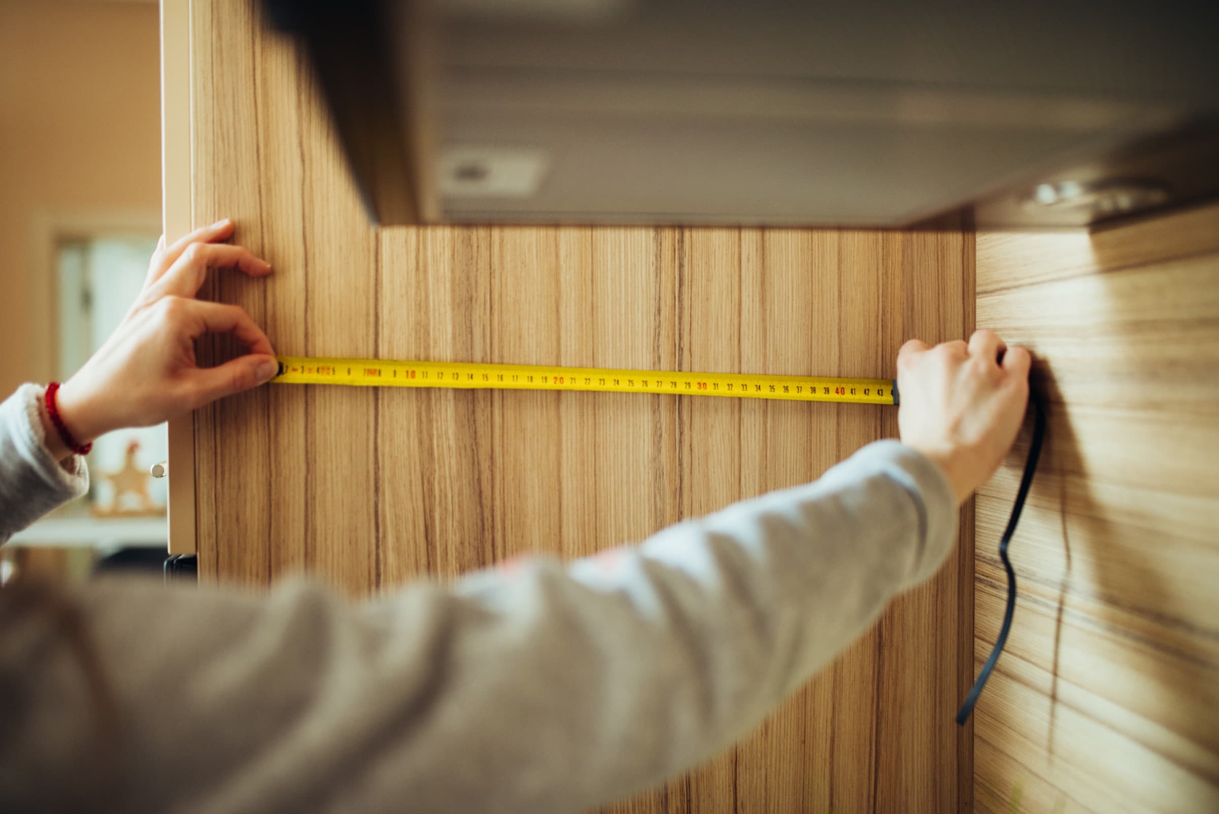Two hands stretching a yellow tape measure across a wood cabinet panel.